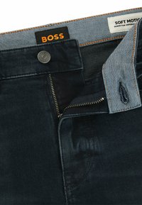 Dark denim jeans with a zipper fly, metal button, and contrasting blue waistband. Features subtle wear patterns and orange stitching.