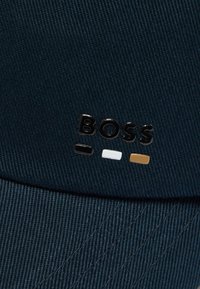 Tumed sinine kangas, millel on ülespoole tõstetud mustad tähed, mis moodustavad sõna "BOSS" ning allpool väikesed mustad, valged ja beežid ristkülikukujulised kujundid.