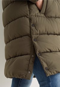 Olivengrøn puffertjakke med quiltet design, der har sorte trykknapper og en høj krave, delvist knappet op, kombineret med blå jeans.