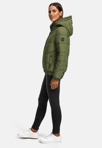 Olijfgroene pufferjack met een capuchon, voorzien van een gequilt ontwerp, twee knopen aan de voorkant en een logopatch op de linkerarm. Gecombineerd met zwarte jeans en witte schoenen.