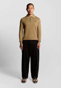 Khaki långärmad polo med tre knappar; en broderad logotyp på bröstet. Parat med lösa svarta byxor och mönstrade bruna sneakers.