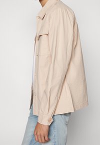 Chaqueta beige claro, texturizada, con un cierre de botones, dos bolsillos en el pecho y puños ajustables. Llevada sobre una camisa blanca y unos jeans azules.