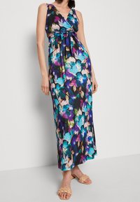 Robe maxi florale noire avec des fleurs bleues, violettes et roses. Décolleté en V, sans manches, taille cintrée avec un lien. Portée avec des sandales beiges.