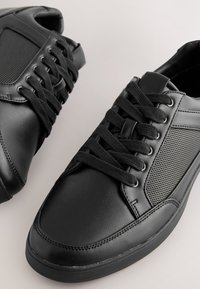 Sneakers in pelle nera con pannelli laterali in mesh, punta tonda, superficie testurizzata e lacci neri. Suola piatta con battistrada minimo.