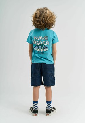 Tricou turcoaz cu grafică "WAVE RIDER", asortat cu pantaloni scurți cargo din denim închis. Adidași negri cu tălpi albe și șosete pătrate albastre.