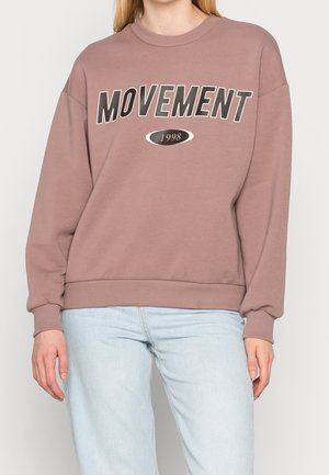 Personne portant un sweat mauve avec le texte "MOVEMENT 1998" et un jean bleu clair, présenté du cou à mi-cuisse sur un fond uni.
