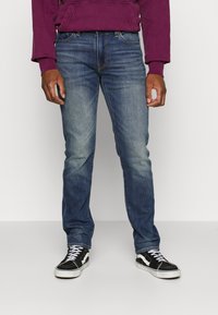 Jean en denim bleu foncé avec une texture légèrement délavée, coupe slim, design à cinq poches, associé à des baskets noires et blanches.