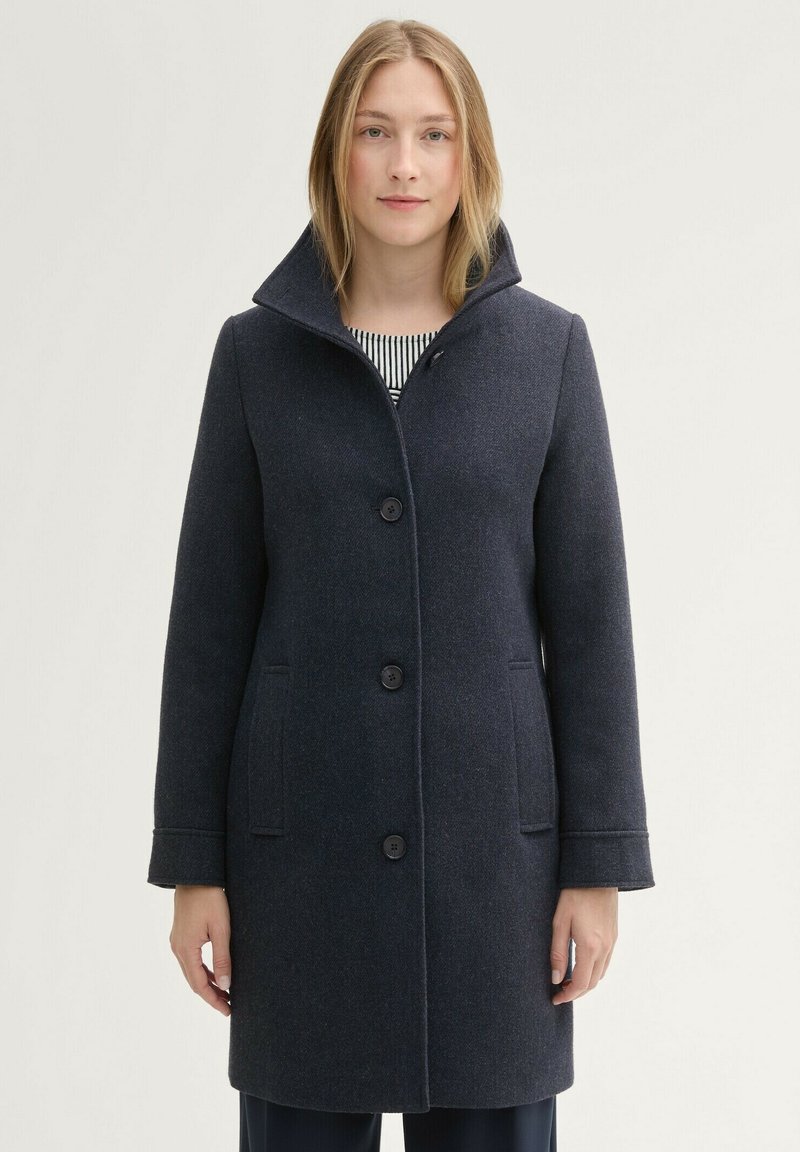 Gyapjú keverék kabát sötét navy színben, magas gallérral, öt gombbal, oldalsó zsebekkel és testre szabott fazonnal. Könnyű anyag.