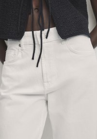 Pantaloni in denim bianco con una texture liscia e design classico a cinque tasche. Abbinati a un top lavorato a maglia blu navy con coulisse.