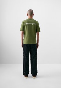 Olivengrøn bomuld t-shirt med korte ærmer og hvid "snow peak" logo på ryggen. Sammen med løstsiddende sorte bukser og brune sko.