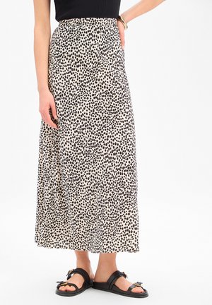 Beżowa spódnica midi w nieregularne czarne grochy, noszona z czarnymi sandałami i czarną bluzką, ręka modelki oparta na biodrze.