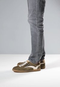 Vihreät mokkanahkaiset tennarit, joissa on valkoisia yksityiskohtia, pyöreät kärjet, teksturoidut pohjat ja valkoiset nauhat. Yhdistetty harmaisiin slim fit -farkkuihin.