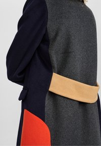 Abrigo de lana con diseño de bloques de color que presenta secciones en azul marino, gris y naranja. Incluye un cinturón beige y un acabado texturizado.