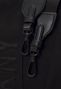 Dettaglio di una borsa nera in tela con accenti in pelle, clip a moschettone e superficie testurizzata con logo in rilievo. Design compatto, hardware funzionale.