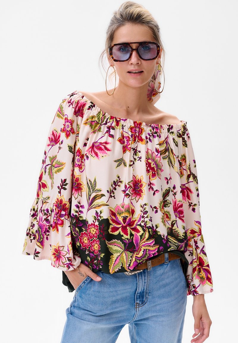 Off-Shoulder-Bluse mit Blumenmuster in rosa und grünen Farbtönen, fließenden Ärmeln und weichem Stoff. Kombiniert mit hellen Jeans.