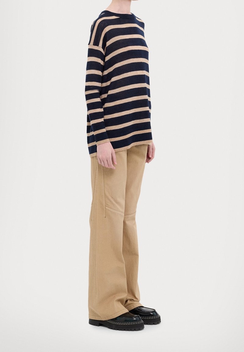 Person in einem navy- und beigen gestreiften Langarmpullover, beigen weiten Hosen und schwarzen klobigen Schuhen, die vor einem weißen Hintergrund steht.