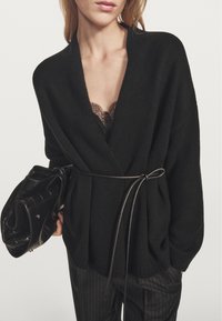 Černý pletený kardigan s hlubokým výstřihem do V, zavazovaný v pase. Má texturovanou látku a pod spodní vrstvou je černá krajka.