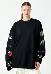 Černý oversized svetr s různými vyšívanými vzory na rukávech, včetně srdcí, slunce a textu, s žebrovým výstřihem a manžetami.