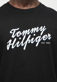 Μαύρο μπλουζάκι με άσπρο και ανοιχτό μπλε σχέδιο της λέξης "Tommy Hilfiger" και την επιγραφή "EST. 1985" στο στήθος.