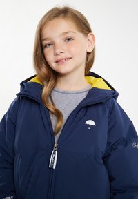Giacca impermeabile blu navy con fodera gialla, zip frontale e piccolo logo di ombrello bianco sul petto. Materiale morbido e imbottito.