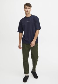 Marine t-skjorte i bomull med korte ermer og broderte tekst, kombinert med grønne tapered bukser og svarte sko. Enkel, uformell design.