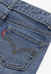 Zadnja džepna stranica plavih traperica s kontrastnim narančastim šavovima i malom crvenom tkaninom oznakom branda s lijeve strane.