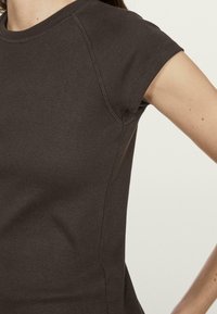 T-shirt marrone a maniche corte con design aderente, texture liscia e cuciture a contrasto. Il tessuto appare morbido, con scollo rotondo.