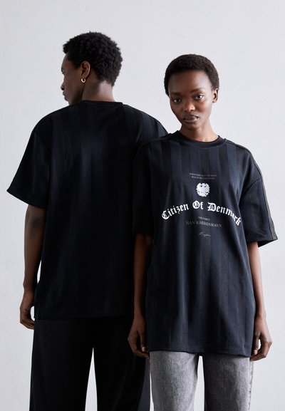 Sort oversized t-shirt med lodret stribemønster, der har hvid tekst og logo på forsiden. Modellen bærer grå bukser og står ryg mod ryg.