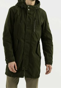 Chaqueta larga verde oliva con capucha, cierres de cremallera y botones a presión, bolsillos laterales y cuerdas ajustables. Hecha de una tela texturizada.