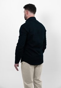 Donkerblauwe overhemd met lange mouwen, kraag en knoopmanchetten, van achteren weergegeven, gecombineerd met lichte broek. Gladde stofstructuur.