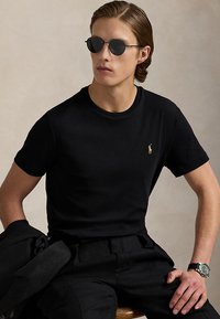 Ung mand iført sorte runde solbriller, sort t-shirt med lille logo, sorte bukser, holder sort jakke og armbåndsur, siddende mod neutral baggrund.