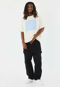 Camiseta de gran tamaño color crema con diseño gráfico azul; pantalones cargo negros con bolsillos de parche; zapatillas blancas. Estilo urbano informal.