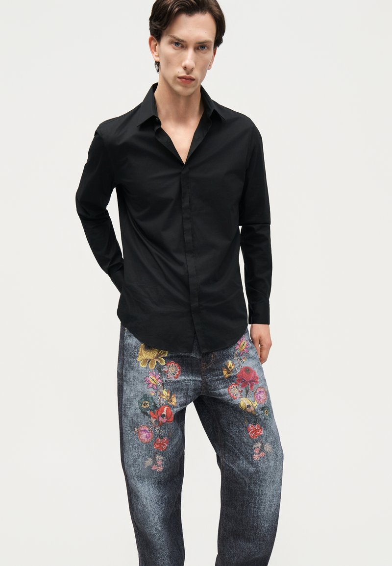 Homme portant une chemise noire à manches longues et un jean bleu avec une broderie florale colorée, debout devant un fond clair uni.