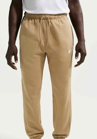 Pantalones deportivos Nike color beige con cintura elástica, cordón ajustable, bolsillos laterales y un logo blanco en el muslo izquierdo. Textura de tela suave.