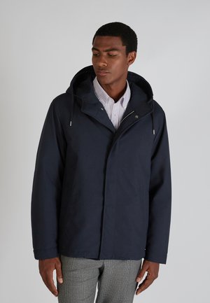 Lichtgewicht marineblau jack met capuchon, voorzien van een rits en zakken aan de voorkant, gecombineerd met een licht gestreept shirt eronder.