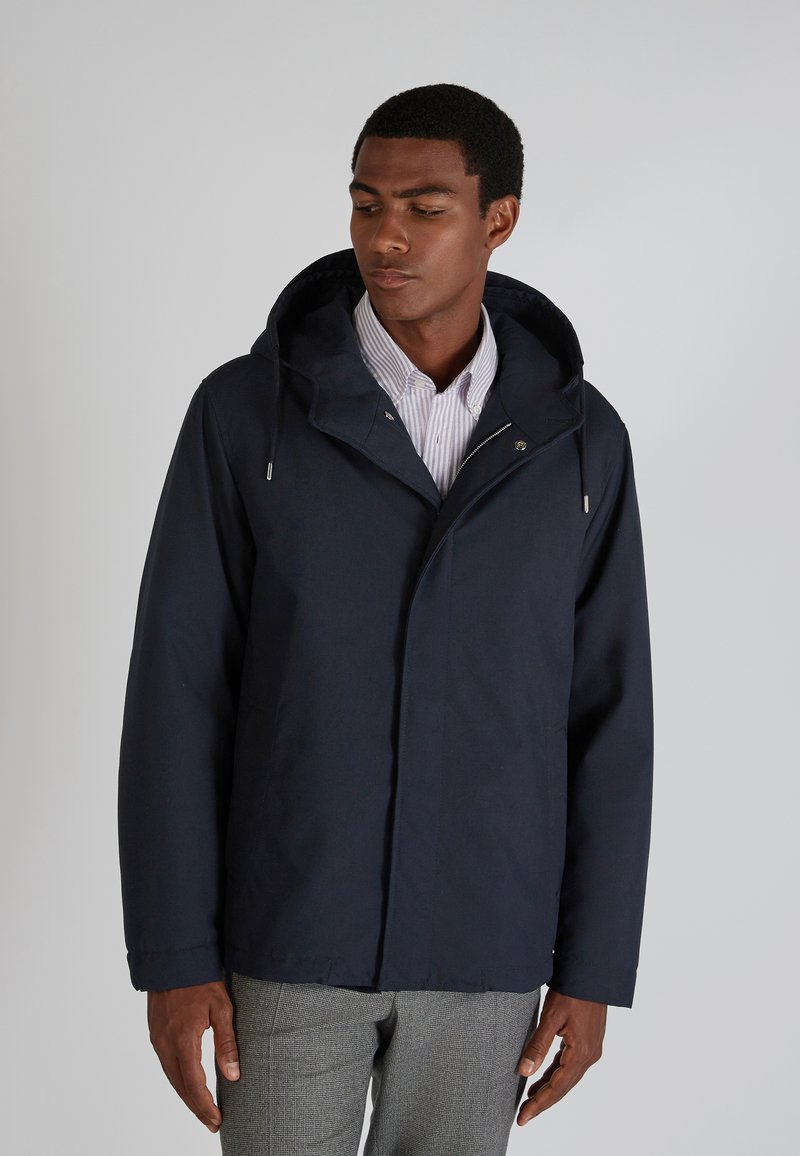 Navy blue lightweight jacket with a hood, featuring a zip closure and front pockets, paired with a light striped shirt underneath.