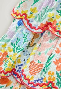 Biele tkanivo s farebnými kvetinovými potlačami v žltej, oranžovej, modrej a zelenej farbe, s červeným vlnitým lemom pozdĺž okrajov.