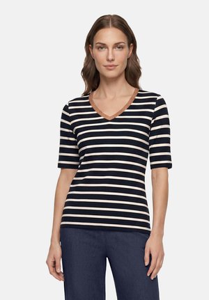 Femme portant un t-shirt à rayures horizontales bleu marine et blanc avec col en V et bordure marron, ainsi qu'un pantalon bleu foncé, debout les bras détendus.