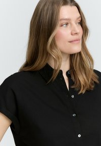 Chemise noire à manches courtes avec un design à col, dotée de boutons sur le devant et d'une texture lisse.