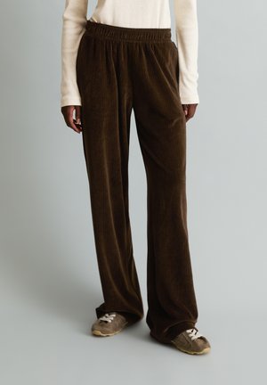 Person wearing loose dark brown corduroy pants with elastic waistband and gray sneakers with white laces against a plain background.