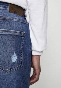 Vaqueros de denim con un acabado azul desgastado, que presentan un parche de cuero marrón en la cintura y un bolsillo trasero parcialmente rasgado. Camisa blanca de manga larga visible.