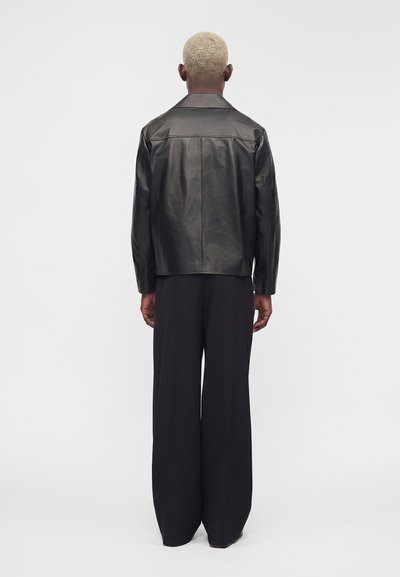 Personne aux cheveux blonds courts portant une veste en cuir noire et un pantalon noir ample, debout de dos sur un fond blanc uni.