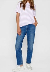 Žena v světle růžovém pruhovaném tričku, modrých džínách a bílých teniskách, stojící s jednou rukou v kapse před jednoduchým pozadím.