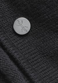 Zwart breiwerk met een gladde textuur, voorzien van een ronde metalen knop met de opdruk "PEARLWOOD." De knop is dicht bij de rand van de stof geplaatst.