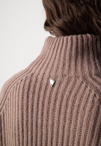 Pull à col roulé côtelé marron avec une petite épingle argentée en forme de cœur dans le dos sous l'encolure, porté par une personne aux cheveux bruns.