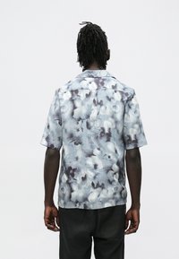 Vista trasera de una persona de piel oscura y rastas, que lleva una camisa de manga corta con un patrón azul grisáceo y pantalones negros, sobre un fondo sencillo.