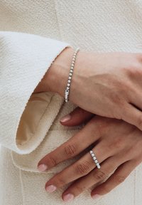 Manos con esmalte de uñas rosa claro que llevan una pulsera y un anillo de cuentas plateadas a juego sobre una manga de tela crema texturizada.