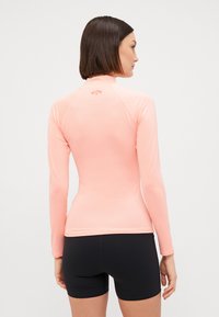 Rash guard a maniche lunghe di colore rosa chiaro, realizzato in tessuto elasticizzato. Presenta un colletto alto e cuciture piatte. Sono visibili anche degli shorts neri.