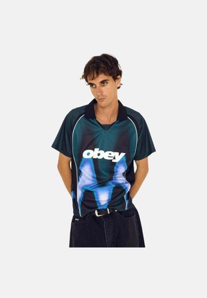 Young man wearing a black and blue short-sleeve polo shirt with "obey" logo, standing with hands behind back against a plain background.