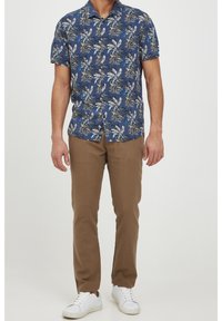 Niebieska koszula z krótkim rękawem z białym i złotym wzorem liści palmowych, zestawiona z brązowymi spodniami i białymi trampkami na neutralnym tle.